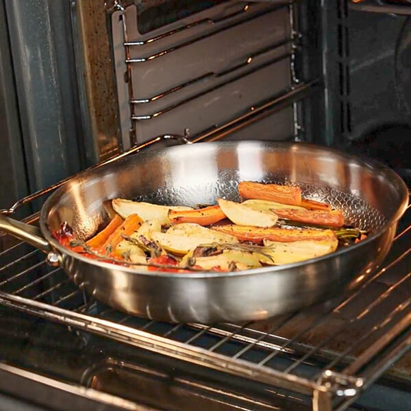 A Titanium steel skillet with a handle sits on an oven rack, cooking sliced potatoes and carrots with vegetables inside an oven.
