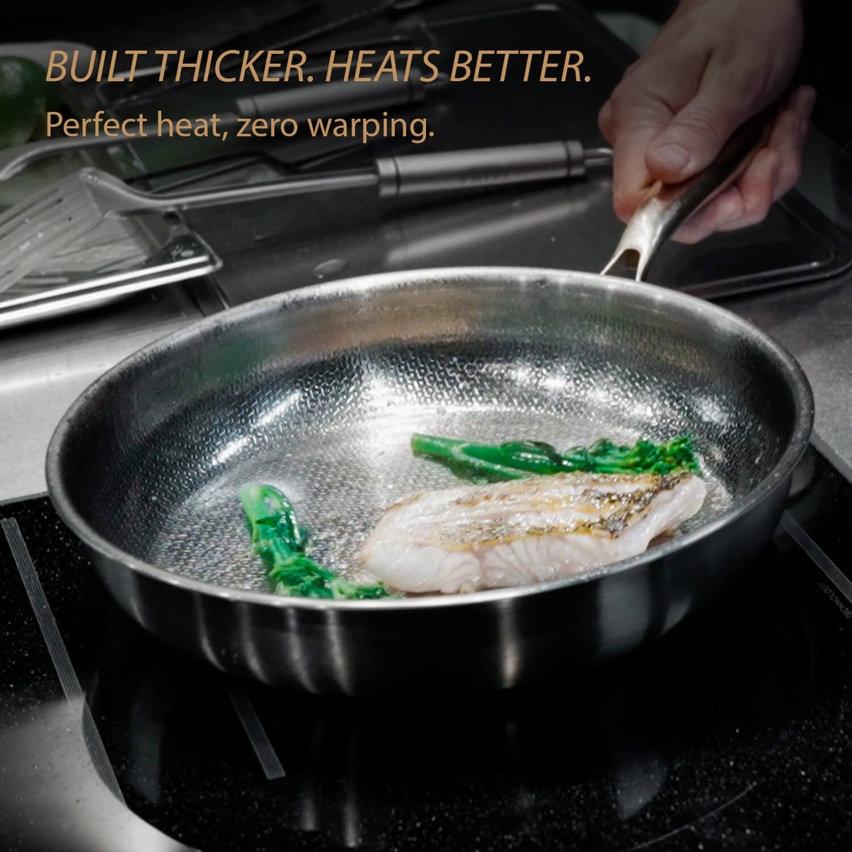 A stainless steel frying pan with a textured interior sits on a stovetop, cooking fish and green vegetables. A hand holds the handle, and promotional text at the top reads 'BUILT THICKER.