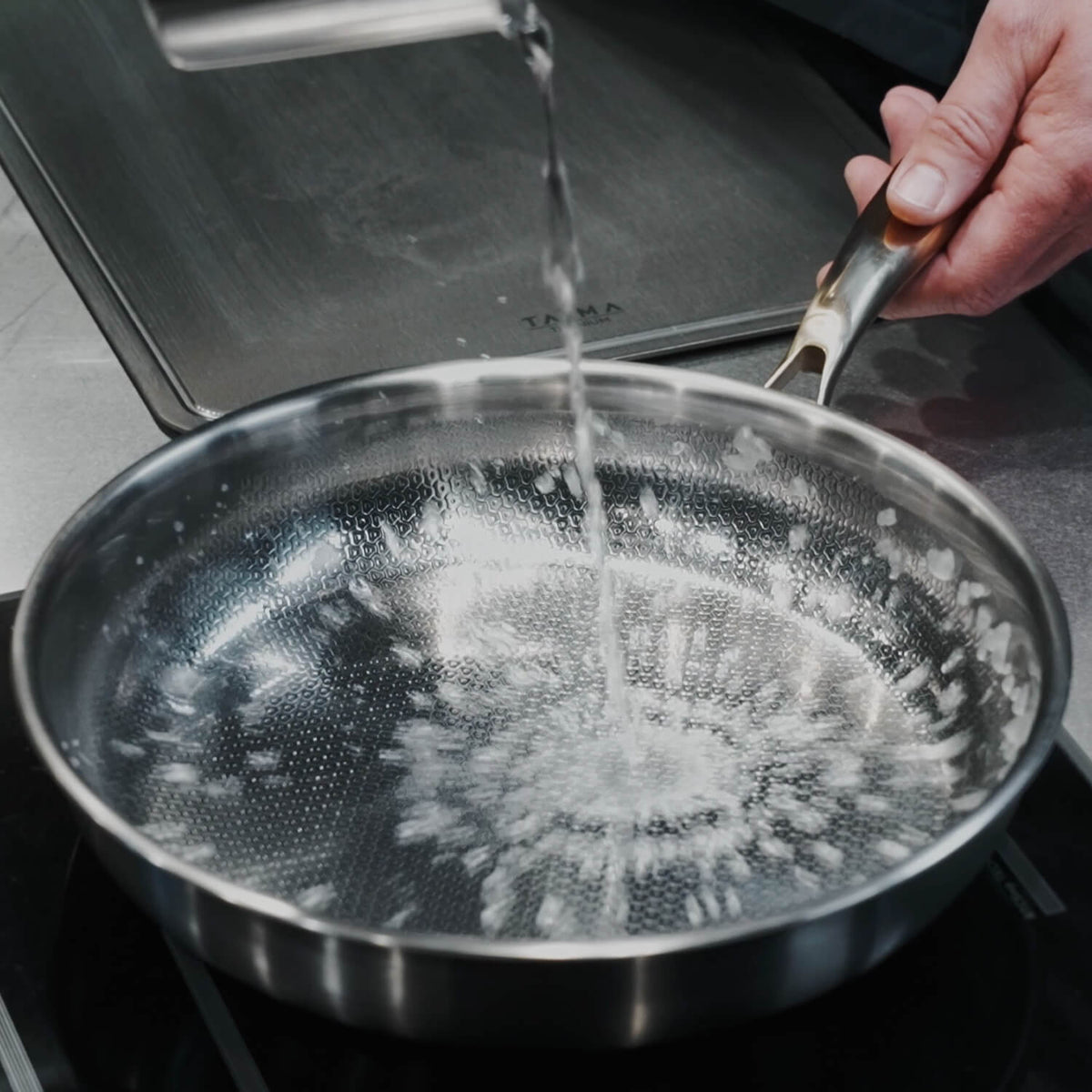 A Titanium frying pan with a textured, dimpled interior surface is shown on a stovetop with water being poured into it. A hand is holding the handle, and a dark stovetop surface is visible in the background.